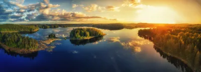 Flygvy över en glittrande sjö med skogsklädda öar och gyllene solnedgång i det svenska inlandet.