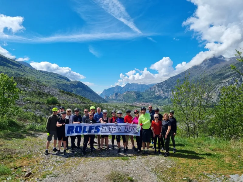 Cykelgrupp från Rolfs Flyg & Buss poserar med banner framför dramatiskt bergslandskap i Italien