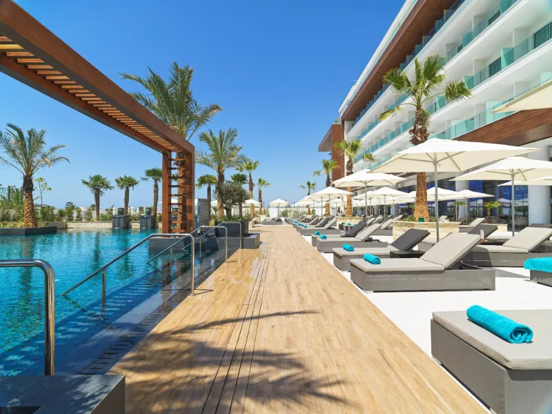 Modernt hotell med pool, solstolar och palmer under en klarblå himmel.