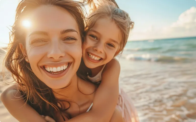 Leende kvinna och barn på en solig strand, där barnet kramar kvinnan bakifrån. Havet och den ljusa sandstranden i bakgrunden skapar en varm och familjevänlig semesterkänsla.