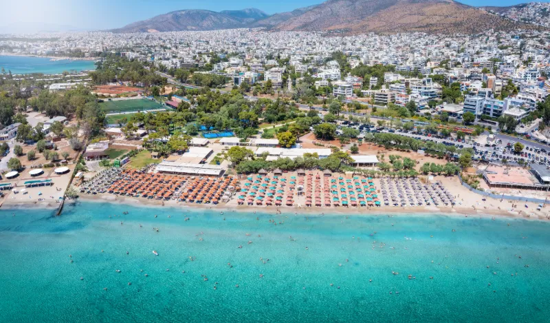 Flygfoto över en lång sandstrand i Aten med kristallklart turkosblått vatten och badgäster som njuter av solen.