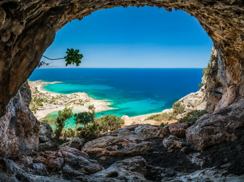 Utsikt över turkost hav och vit sandstrand genom öppningen i en klippgrotta högt ovanför kusten.