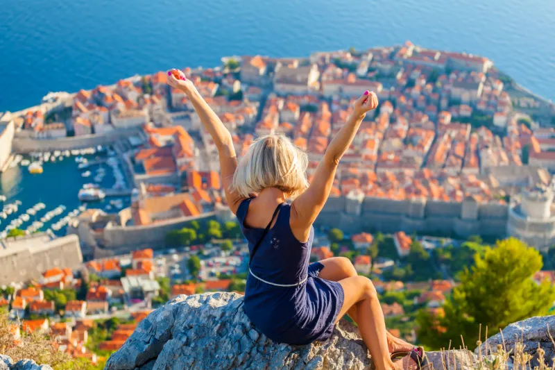 Person sitter på en klippa med armarna uppsträckta och blickar ut över Dubrovniks historiska stadskärna och Adriatiska havet.