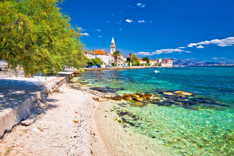 Kustnära stad i Kroatien med kristallklart turkost vatten, stenstrand och historiska byggnader längs strandpromenaden.