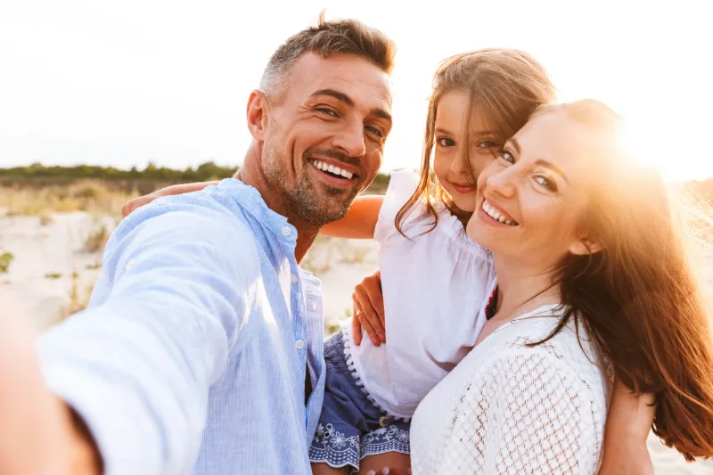 Familj som tar en selfie på en solig strand. Två vuxna och ett barn ler mot kameran medan de står tätt tillsammans i det varma kvällsljuset