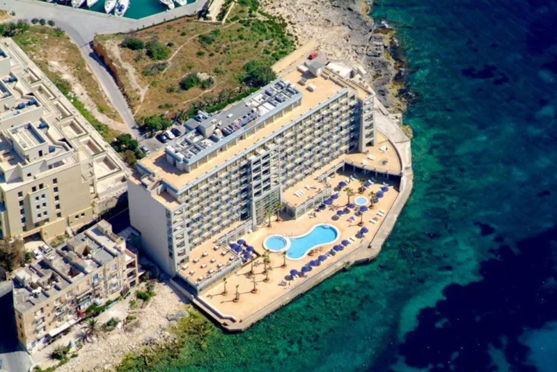 Flygvy över ett kustnära hotell med poolområde och solterrass direkt vid det kristallklara turkosblå havet.