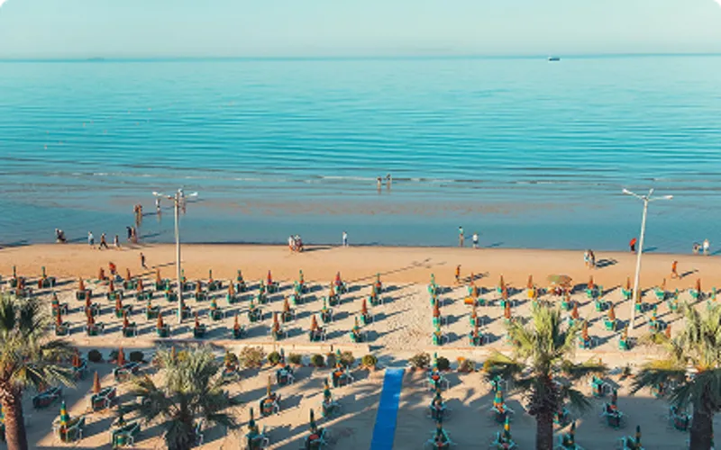 Flygvy över en bred sandstrand med rader av solstolar och parasoller vid ett lugnt, spegelblankt hav. Durres Riviera