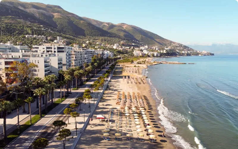 uststad vid Adriatiska havet med lång sandstrand, palmkantad strandpromenad och rader av solstolar längs vattnet. Vlora