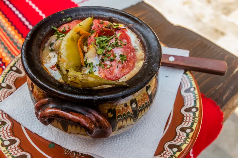Traditionell bulgarisk maträtt serverad i en handgjord lergryta på ett dekorerat fat.