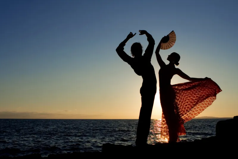 Två flamencodansare i silhuett vid havet under en färgstark solnedgång.