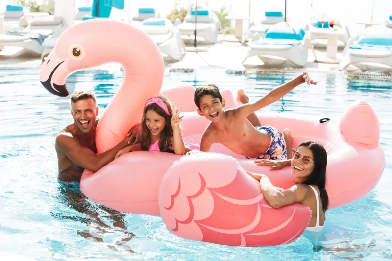 Alt-text: En glad familj leker i en hotellpool på en stor rosa flamingoflotte. Två vuxna och två barn skrattar och poserar medan de flyter i det klara vattnet.