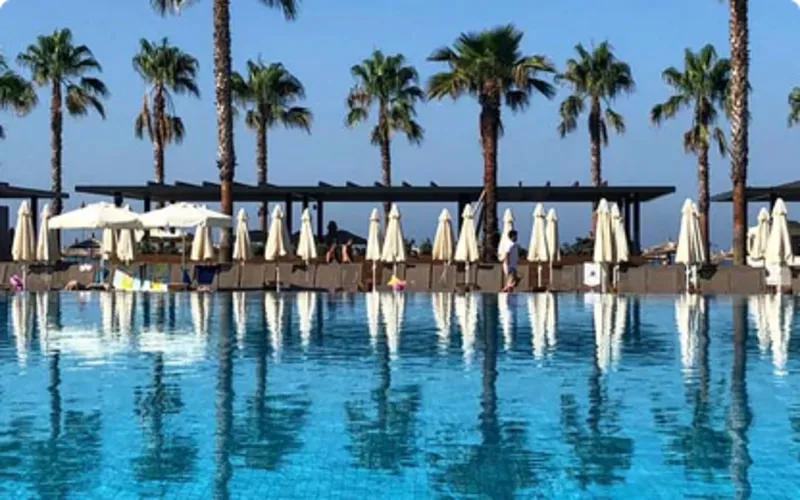 Stor hotellpool med klart blått vatten i förgrunden. Runt poolen står vita parasoller på rad, och bakom dem syns höga palmer mot en klarblå himmel